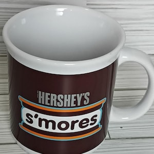 May include: A white ceramic mug with a brown band featuring the Hershey's S'mores logo. The mug has a white interior and a curved handle. The mug is set against a light wood-grain background.