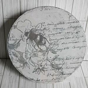 May include: A round, decorative wooden piece with a distressed, vintage design. The piece features a detailed illustration of a bee and floral elements in gray tones, overlaid with script. The edges are lightly distressed.
