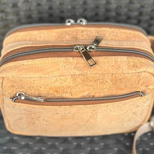 May include: A light brown cork crossbody bag with two zippered compartments and a long adjustable strap. The bag has a textured cork surface and silver zippers.