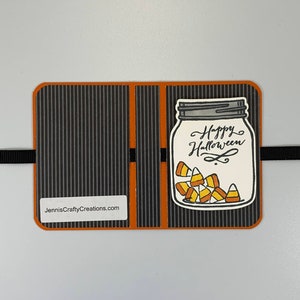 May include: A black and orange card with a striped background. The card has a cut-out image of a mason jar filled with candy corn. The text "Happy Halloween" is written on the jar. The website "JenniesCraftyCreations.com" is printed on the card.