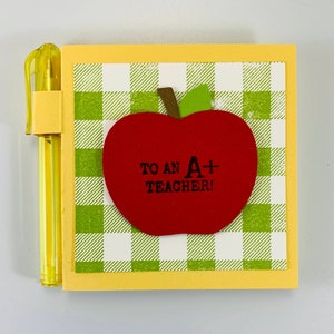May include: A yellow notepad with a green and white checkered pattern and a red apple with the text "To an A+ Teacher!" on it.