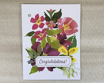 May include: A white card with a watercolor floral design featuring pink, purple, and yellow flowers. The card has a white banner with the text "Congratulations!" printed on it.