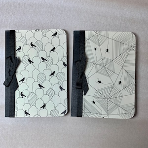 May include: Two black and white patterned notebooks with black ribbon closures. One notebook has a pattern of tombstones and crows, the other has a spiderweb pattern.