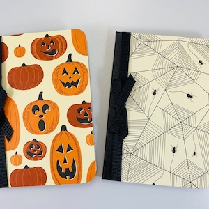 May include: Two Halloween-themed notebooks with black ribbon closures. One notebook features a pattern of orange pumpkins with jack-o'-lantern faces. The other notebook features a pattern of black spiderwebs on a cream background.