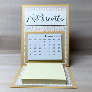 May include: A yellow and gray desk calendar with a sticky note pad. The calendar is for January 2020 and has the words "just breathe." printed on the top.