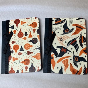 May include: Two small notebooks with black ribbon closures. One notebook has a white background with a repeating pattern of brown and orange potion bottles. The other notebook has a white background with a repeating pattern of black witches' hats.