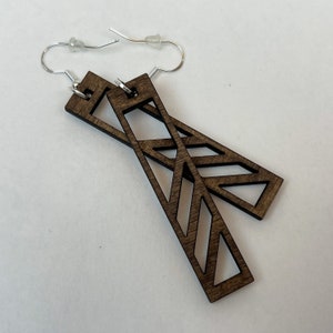 Puede incluir: Un par de pendientes de madera marrón con un diseño geométrico. Los pendientes tienen forma de rectángulos largos con recortes en forma de triángulos y zigzags.