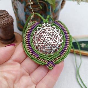May include: A handmade macrame pendant necklace featuring a silver-colored geometric design at the center. The pendant is surrounded by green, purple, and tan macrame knots. The necklace is a unique piece of jewelry.