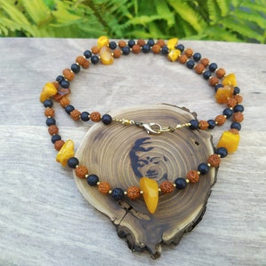 Puede incluir: Un collar con cuentas de ámbar y marrón oscuro, intercaladas con cuentas negras más pequeñas. El collar se exhibe sobre una superficie de madera con un diseño tallado. El cierre es de color dorado.