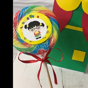 May include: A large, colorful lollipop with a cartoon character label, wrapped in clear cellophane. The lollipop has a red ribbon tied to the wooden stick. A green, red, and yellow paper decoration is in the background.