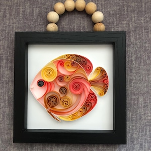 Op de afbeelding: Een ingelijst papieren quilling-kunstwerk van een rode, oranje en gele vis. De vis is gemaakt van gekruld papier en is geplaatst tegen een witte achtergrond. De lijst is zwart en heeft een handvat van houten kralen.
