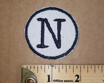 May include: A circular patch with a white background and a black border. The letter "N" is embroidered in black within the circle. A wooden ruler with inch measurements is next to the patch.