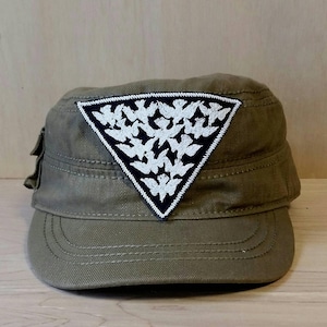 May include: Olive green military-style cap with a triangular patch. The patch features a black background with a white dove design. The cap has a short brim and a structured crown, suitable for casual wear.