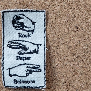 May include: A rectangular white fabric patch with black embroidered illustrations of the hand gestures for Rock, Paper, and Scissors. The words "Rock", "Paper", and "Scissors" are embroidered below each respective hand gesture.