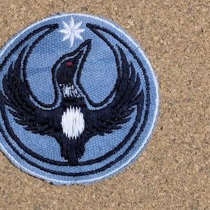May include: Embroidered patch showing a black bird with spread wings, a white chest, and a red eye, on a light blue background. A white star is above the bird's head, with a blue border around the patch.