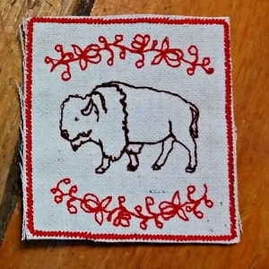 May include: Square, off-white fabric patch with a hand-stitched image of a bison in black thread. The patch is bordered with red stitching and features red floral embroidery along the top and bottom edges. The bison is facing left.
