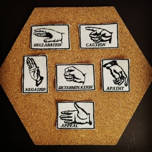 May include: A collection of seven white patches with black embroidered hand gestures and text. The patches are arranged on a corkboard. Each patch features a different hand gesture with a corresponding word: Declaration, Caution, Negation, Determination, Apathy, and Appeal.