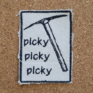 May include: A rectangular patch with a white background and a black border. The patch features a black ice axe illustration and the word "picky" repeated three times in black, stylized text. The patch is likely made of fabric.
