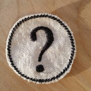 May include: A circular patch with a white fabric base and a black embroidered border. The patch features a large black question mark in the centre. The patch is set against a light brown wooden surface.