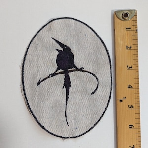 May include: An oval patch featuring a black bird silhouette perched on a branch. The bird has a long tail and is set against a light gray background. A wooden ruler is next to the patch.