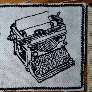 May include: Embroidered patch with a black and white illustration of a vintage typewriter. The design is stitched on a square, off-white fabric with a black border. The patch is approximately 5 cm wide.