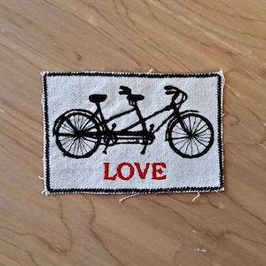 Peut inclure: Un écusson rectangulaire avec une illustration d'un tandem noir sur fond blanc. Le mot "LOVE" est en lettres rouges sous le vélo. L'écusson a une bordure noire cousue.