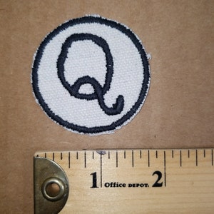May include: A circular patch with a white background and a black embroidered outline. The letter "Q" is embroidered in black within the circle. A wooden ruler with inch markings is next to the patch.
