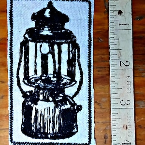 May include: A black and white patch featuring a vintage lantern design. The patch is rectangular with a stitched border. A ruler is next to the patch, showing measurements in inches.