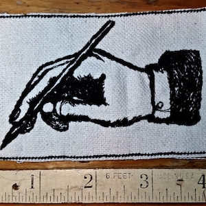 May include: A rectangular patch featuring a black and white illustration of a hand holding a pen. The patch is made of a textured, off-white fabric with a black stitched border. A ruler is placed below the patch.
