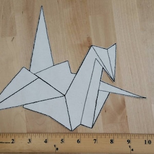 May include: A white origami crane with black outline details. The paper bird is folded into a traditional crane shape, with wings and a pointed beak. The crane is placed on a wooden surface, with a ruler showing inches.