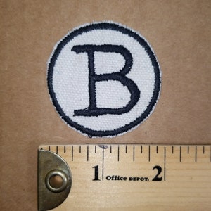 May include: Circular white patch with a black embroidered border and a large black letter "B" in the center. A wooden ruler with black markings and the text "Office Depot" is next to the patch.