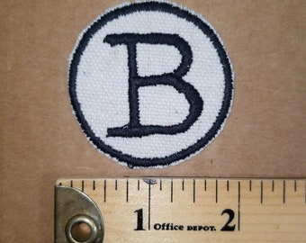 May include: Circular white patch with a black embroidered border and a large black letter "B" in the centre. A wooden ruler with black markings and the text "Office Depot" is next to the patch.