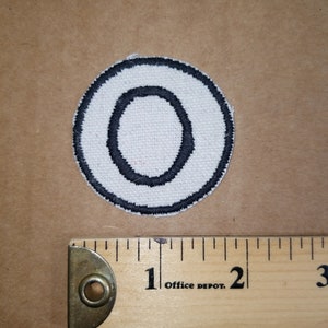 Peut inclure: Écusson blanc circulaire avec un contour noir et un cercle intérieur noir. Le patch est placé sur une surface marron à côté d'une règle en bois. La règle est marquée en pouces et porte le texte "Office Depot".