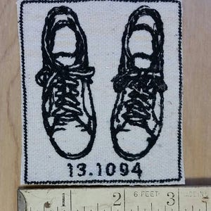 May include: Embroidered patch featuring a pair of black sneakers. The sneakers are stitched in black thread on a cream-colored fabric. The date "13.10.94" is embroidered below the shoes. The patch is approximately 2.5 inches wide.