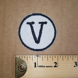 May include: A circular patch with a white background and a black border. The letter "V" is embroidered in black in the centre. A wooden ruler with "Office Depot" printed on it is next to the patch.