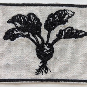 Puede incluir: Un parche cuadrado de color blanquecino con una imagen bordada en negro de una planta de remolacha. La remolacha tiene una raíz redonda, hojas verdes y un sistema de raíces detallado. El parche está enmarcado por un borde negro.