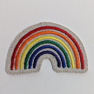 May include: Embroidered rainbow patch featuring a vibrant arc of red, orange, yellow, green, blue, and indigo threads. The rainbow design is stitched onto a light beige fabric, creating a colorful and cheerful accessory.