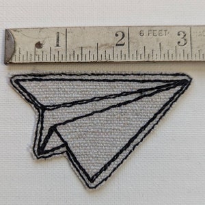 May include: A grey fabric paper aeroplane patch with black stitched outline. A vintage measuring tape is placed above the patch, showing measurements in feet and inches.