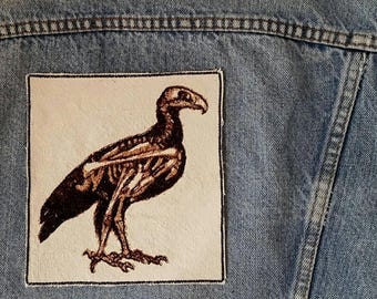Könnte beinhalten: Ein quadratischer Aufnäher mit einer detaillierten Illustration eines Vogelskeletts in Braun- und Beigetönen. Der Aufnäher ist auf eine hellblaue Jeansjacke genäht, wodurch die Kunstwerke vom Stoff abgesetzt werden.