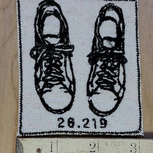 May include: Embroidered patch featuring a pair of black sneakers, stitched on a white fabric square with a black border. The number "26.219" is printed below the shoes. The patch is approximately 2 inches wide.
