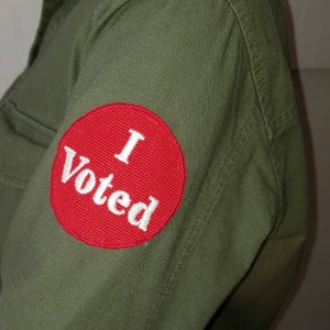 May include: Olive green shirt with a red circular patch that reads "I Voted" in white text. The patch is on the left sleeve of the shirt. The shirt has a textured appearance.