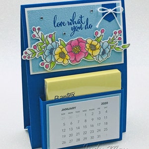 Puede incluir: Un organizador de escritorio azul con un porta notas adhesivas amarillo y un calendario para enero de 2020. La parte delantera del organizador tiene un fondo azul con el texto "love what you do" y un diseño floral.