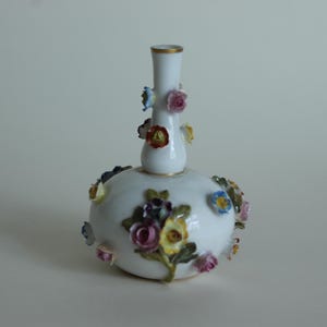 Può includere: Un vaso in porcellana bianca con base bulbosa e collo sottile, decorato con fiori in ceramica tridimensionali applicati nei toni del rosa, giallo, blu e viola, con bordi dipinti in oro sulla parte superiore e sulla base.