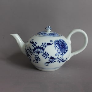 Könnte beinhalten: Eine weiße Porzellanteekanne mit einem blauen Blumenmuster aus filigranen Ranken und Blüten. Die Teekanne verfügt über einen geschwungenen Henkel, einen spitz zulaufenden Ausguss und einen passenden Deckel mit einem kleinen, verzierten Knauf.