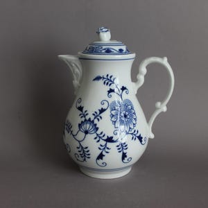 May include: A white porcelain coffee pot featuring a blue onion pattern with floral and vine motifs. The pot includes a matching lid with a small rose-shaped finial, a curved handle, and a spout with decorative molded details at the base.