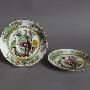 May include: Two white porcelain saucers with a gold rim and a colourful floral and bird design. The design features a peacock and other birds on a tree branch with flowers.