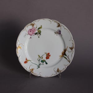 Peut inclure: Une assiette en porcelaine blanche avec un bord doré et des motifs floraux dans des tons de rose, rouge, jaune et vert.