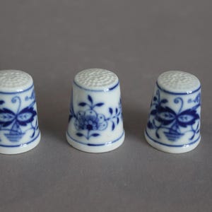 Könnte beinhalten: Drei weiße Fingerhüte aus Keramik in einer Reihe, jeder mit einem blauen Blumenmuster und einer strukturierten, genoppten Oberseite. Die Fingerhüte haben eine glatte, glänzende Oberfläche und eine zylindrische Form mit einem leicht ausgestellten Rand.