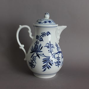 May include: A white porcelain coffee pot featuring a blue floral pattern. The pot has a curved handle, a spout, and a removable lid with a small knob. The blue design consists of intricate botanical illustrations, including leaves and flowers, painted on the exterior surface of the vessel.