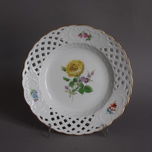 May include: A white porcelain plate with a lattice design around the rim and a gold edge. The center features a yellow rose with green leaves and purple and blue flowers. A decorative piece for serving.